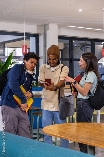 Diverse students leaning in, showing red phone in campus lounge, holding notebooks and backpacks