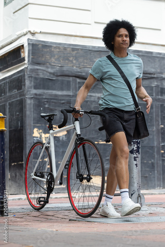 Foto African-American man walking white bike red rim on sidewalk wearing black crossb