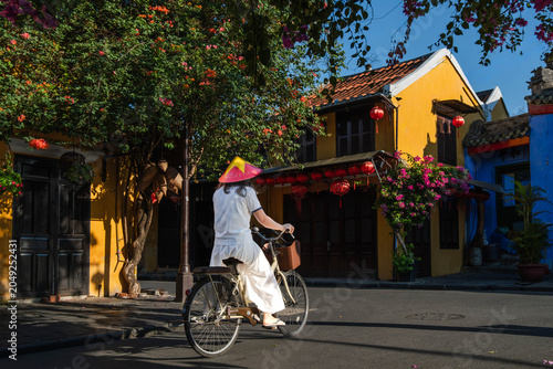 Woman in traditional conical hat riding a bicycle down a historic town street