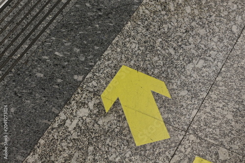 Yellow directional arrow painted on speckled granite floor indicating movement forward