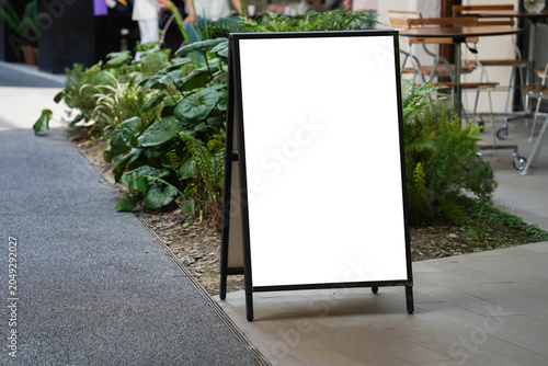 Blank white outdoor advertising stand sandwich board mock up template. Clear street signage board placed by an outdoor dinning area of a restaurant.