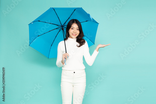 Happy young Asian woman holding a blue umbrella and showing open palm presenting copy space, smiling at camera in white winter outfit isolated over a mint green background