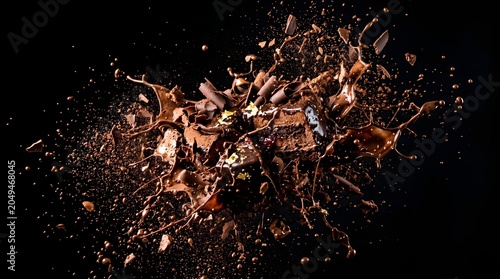 A high speed photography shot capturing a dramatic explosion and splash of liquid chocolate and cocoa chunks frozen mid air against a solid black background.