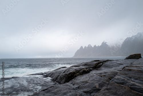 The dramatic 'Devil's Teeth' rock formations at Tungeneset viewpoint on the stormy Norwegian coast with misty mountains in the distance.