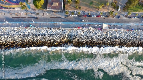 Tableau sur toile High angle drone view of ocean waves breaking against a rocky coastal breakwater beside a waterfront promenade