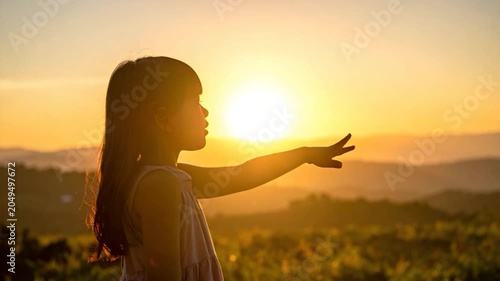 A young girl reaches towards a sunset, silhouetted against the golden sky