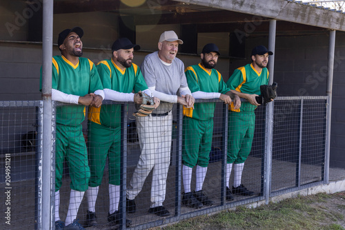 Photography Diverse male teammates leaning on chain-link railing in dugout watching game wit
