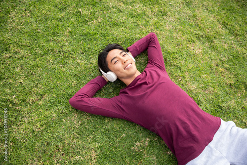 Εκτύπωση καμβά Asian man lying on grassy lawn wearing white headphones and maroon shirt and lig