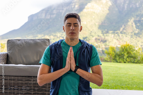 Quadro su tela Man praying, kneeling on patio beside wicker sofa and lawn, wearing teal tee and