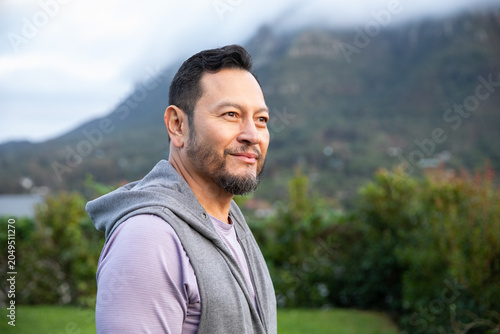 Mature Asian man standing outdoors near shrubs and mountains, wearing grey hoodie over purple shirt