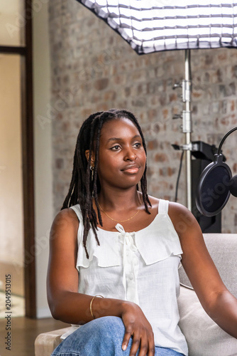 African woman sitting on light-colored sofa in studio recording with microphone and softbox light