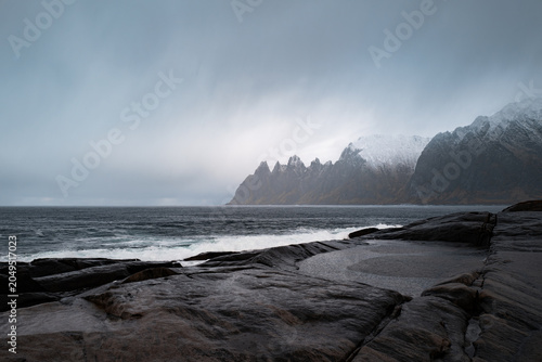 Moody seascape with dark rugged rocks, stormy white-capped waves, and distant snow-covered mountains under a dramatic overcast sky.