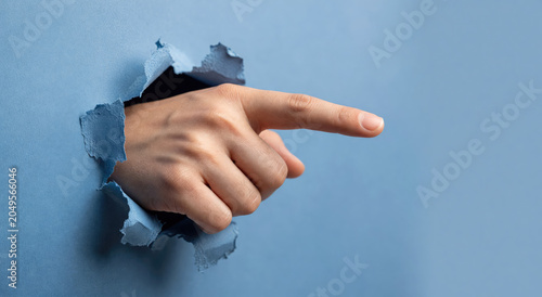 Woman hand pointing finger through a torn blue paper hole. Concept of direction, choice and discovery. Symbolic gesture of communication and information highlighted by a rip in background.