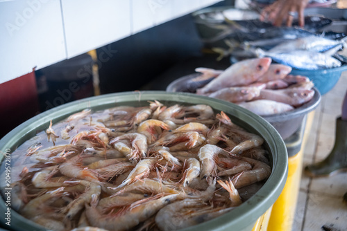 Fresh raw shrimp displayed in a green plastic basin at a traditional seafood market stall with fish blurred in the background.