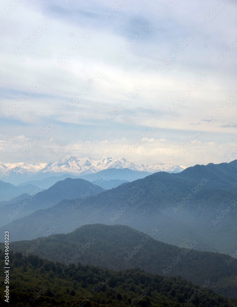 Fototapeta premium Distant snow-capped Caucasus mountain peaks behind misty blue layers of hills under a cloudy sky