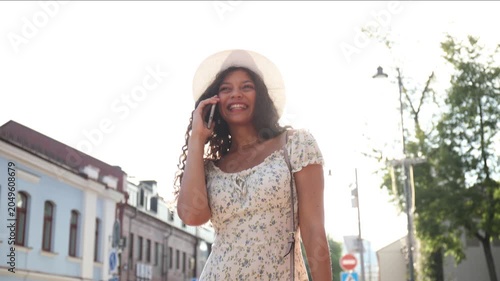 Attractive laughing woman talking on cellphone while walking in summer street in city.