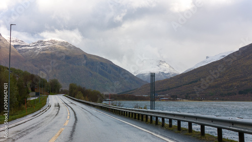 Foto A winding coastal road with guardrails and lamp posts reflected on a mirror-like lake, leading towards misty, snow-capped mountains under a moody overcast sky in Norway
