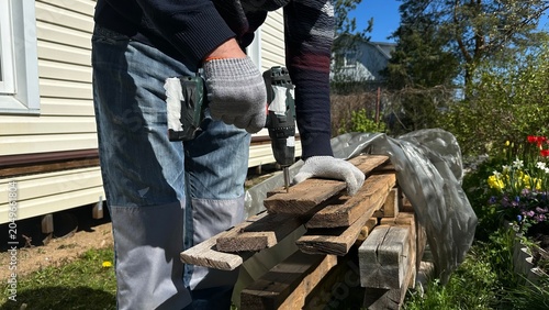 Hands in protective work gloves using a power drill on a wooden surface during home renovation. DIY woodworking, cottage repair, construction, craftsmanship and industrial work.