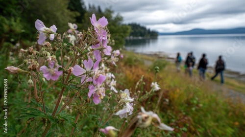 Wildflowers bloom along a peaceful lakeside path, blurred figures stroll onward