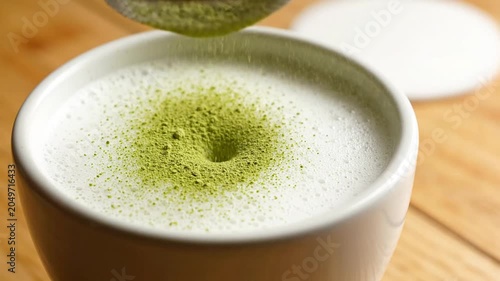 Matcha powder being sifted over a frothy milk latte in a white cup on a wooden table, creating a vibrant green drink