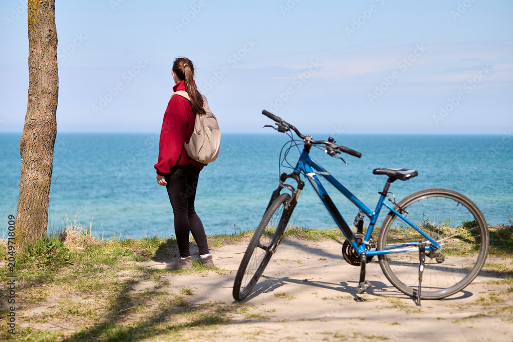 Fototapeta premium Young woman with ponytail standing by ocean shore with blue bicycle on sunny day