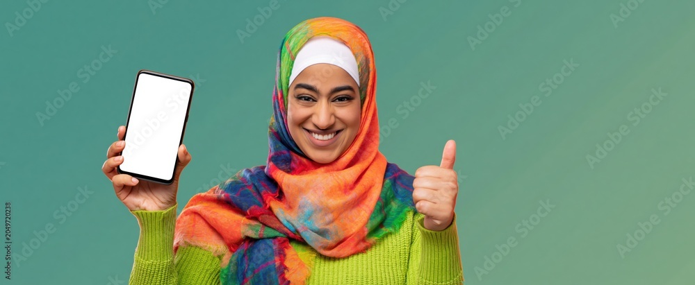Obraz premium Smiling Middle Eastern woman in colorful hijab holds smartphone displaying blank screen and makes thumbs-up gesture.