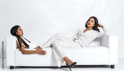 Two women in stylish polka dot suits lounging on a white modern sofa, relaxed pose, indoor studio setting