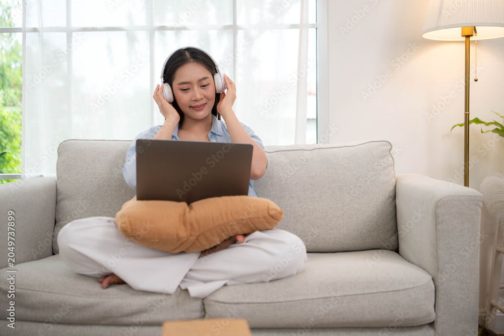 Fototapeta premium Young Asian woman using a laptop and wearing headphones while sitting on a sofa at home