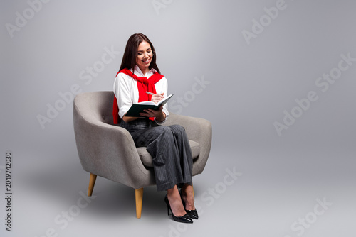 Businesswoman writing in a notebook while seated in a modern armchair  smiling and working casually in a studio with gray background