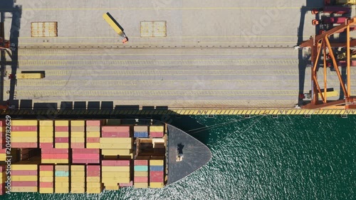 Industrial port scene featuring an aerial top-down view of a massive container cargo ship slowly moving along the pier, showcasing the scale of global trade and maritime logistics operations