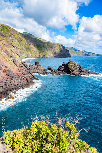 Crashing white waves around black volcanic rock formations under a bright, cloudy sky at Nogales beach, La Palma, Canary Islands, Spain