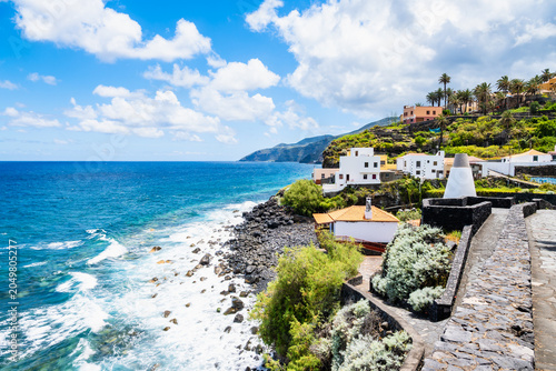Stone promenade overlooks a rocky volcanic shoreline with crashing white waves and white coastal houses in San Andres, La Palma, Canary Islands, Spain