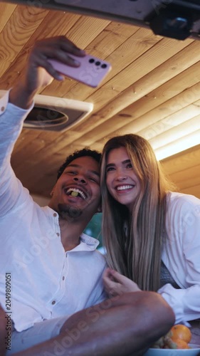 Happy young couple enjoying a holiday inside their camper van. The woman playfully feeds her boyfriend while he takes a selfie with a smartphone