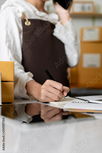 Entrepreneur writing customer order details in notebook near cardboard parcel boxes during online shop logistics process