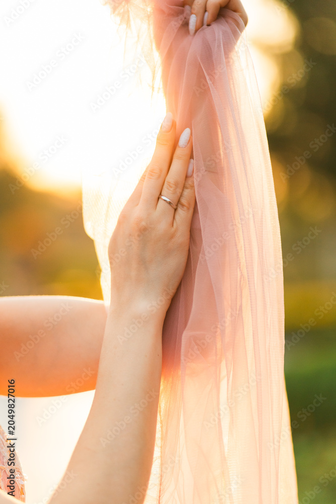 Naklejka premium Young woman's delicate hands with a nice manicure and wedding ring holding a flowing pink tulle scarf at sunset.