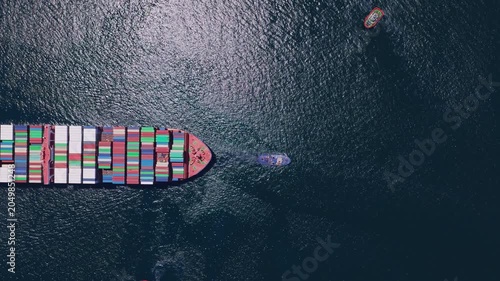 Majestic aerial top view of a large container ship cruising on the open sea, assisted by two powerful tugboats during its voyage, symbolizing global trade and logistics industry