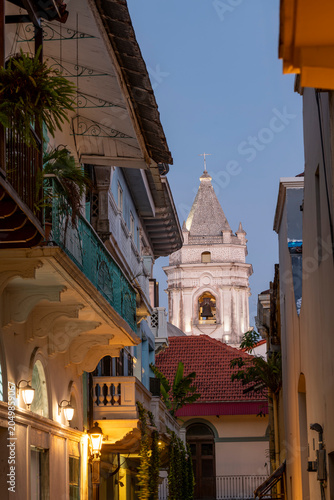 Buildings with colonial architecture lining a narrow street in casco viejo, Panama city, converge towards the iconic bell tower of the Cathedral basilica of st. Mary under the evening sky.
