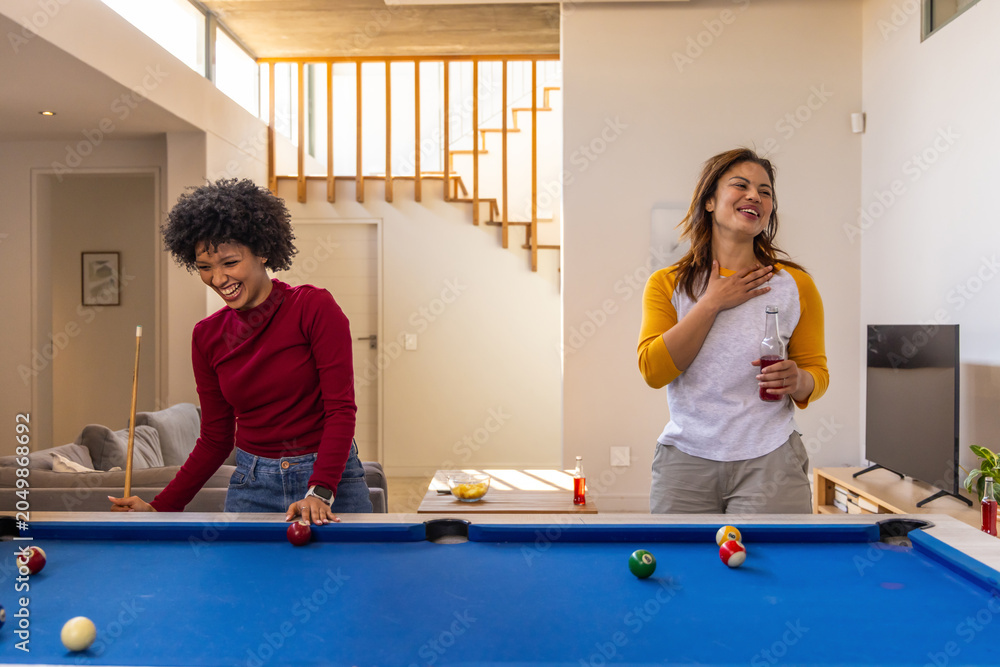 Obraz premium Diverse female friends laughing and playing pool at home with cue stick, holding beverage