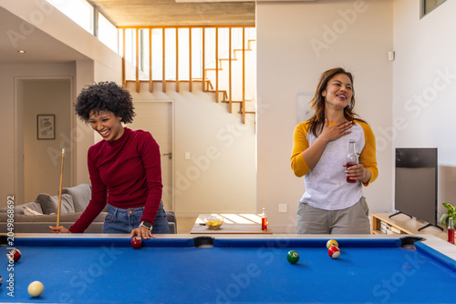 Diverse female friends laughing and playing pool at home with cue stick, holding beverage