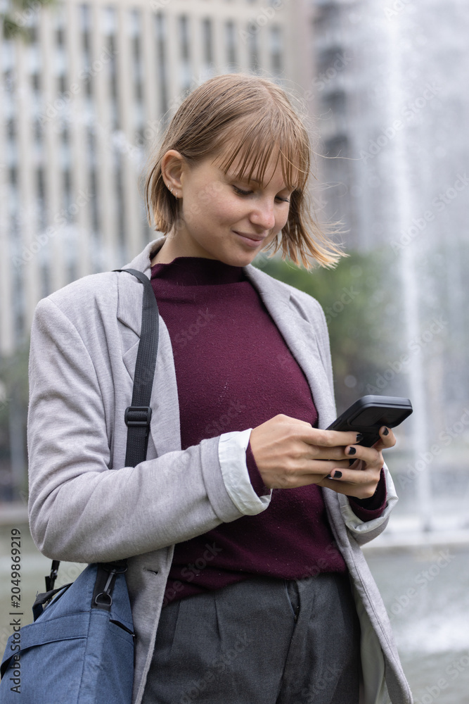 Obraz premium Adult woman standing in plaza by fountain holding smartphone wearing blazer and blue crossbody bag