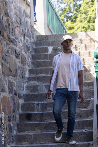 African American man walking down urban stone steps by green handrail, wearing cap, litter on step