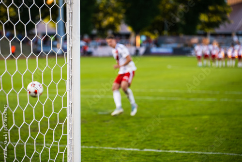 Schilderij op canvas A soccer player in a white uniform kicking a penalty shot towards the goal during a football match, viewed through the net