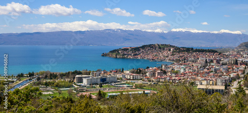 Photography An expansive panorama showcases Ohrid's lively cityscape sprawling along the deep blue lake, framed by distant snow-covered peaks under a partly cloudy spring sky