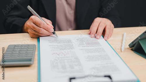 Εκτύπωση καμβά Professional business person wearing dark blazer sits wooden desk reviewing financial document clipboard holding silver pen near calculator tablet stylus workday office setting indoors this afternoon