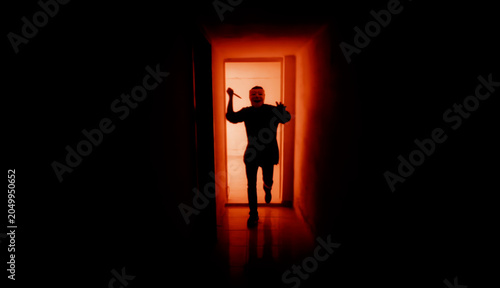 Cuadro en lienzo Masked figure silhouetted in doorway holding knife in dark corridor with dramatic orange backlight