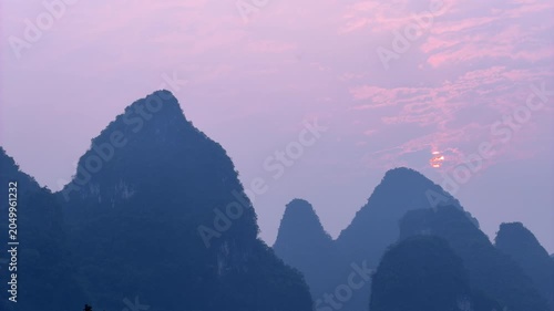 Timelapse of a beautiful pink and purple sunset, with the sun slowly setting behind the dramatic silhouettes of karst mountain peaks in Yangshuo, Guilin, China. Atmospheric evening twilight and golden
