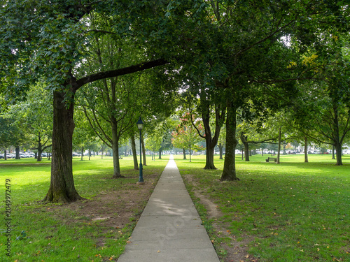 A paved path cuts through the center green of downtown Guilford, Connecticut (CT) shaded by mature trees.