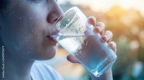 A close up of a sweating person drinking a refreshing glass of cold water during hot summer weather.