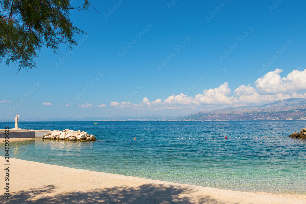 Fototapeta premium Scenic coastal view of Postira village on Brac Island, Croatia. Crystal clear turquoise water of the Adriatic Sea, rocky beach, and distant mountains on the horizon under a bright blue summer sky