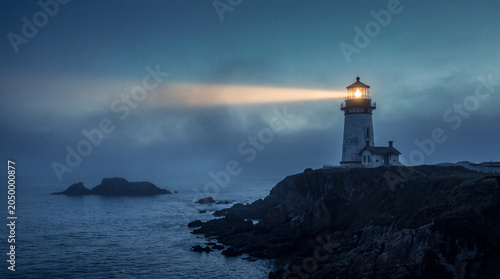 Lighthouse shining a bright beam of light through thick fog over the dark ocean water and rocky coast at night